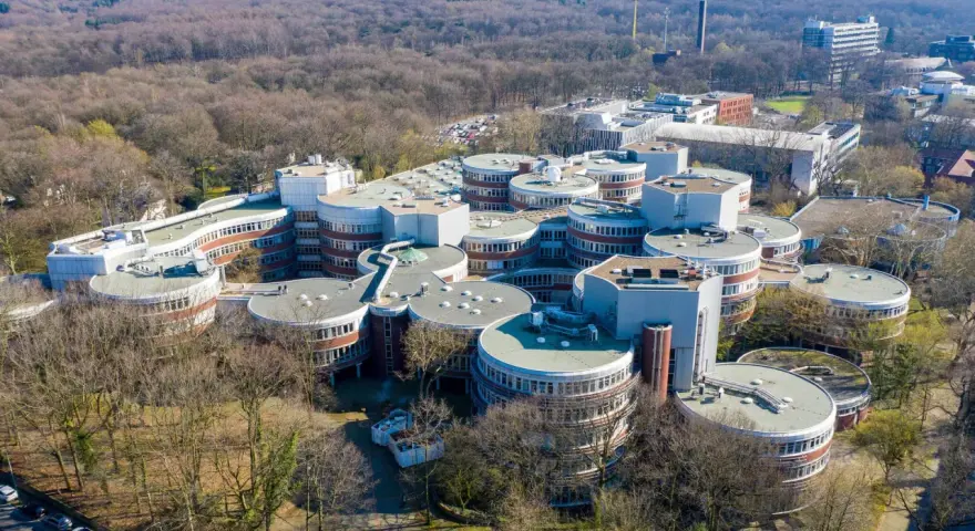 Luftaufnahme des Campus der **Uni Duisburg Essen** mit seinen charakteristischen runden Gebäuden, umgeben von Bäumen.