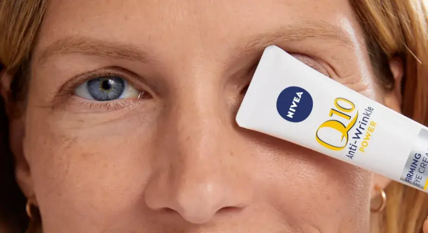 Kobieta z niebieskimi oczami, z kremem Nivea Q10 Anti-Wrinkle Power zakrywającym jedno oko. Skóra wokół oczu ma drobne kurze łapki.