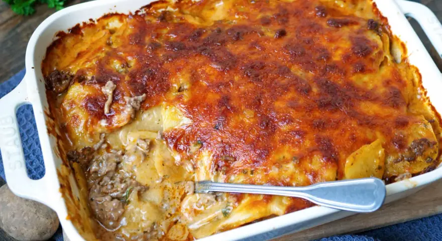 Omas Kartoffelauflauf mit Hackfleisch, goldbraun überbacken, dampft in einer weißen Auflaufform. Eine Gabel steckt in der saftigen Schicht.