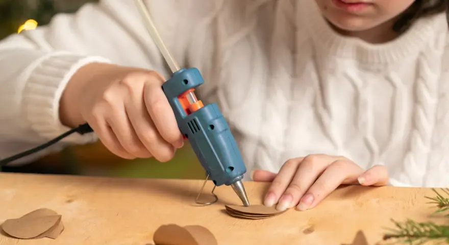 A child uses a hot glue gun to craft heart shapes from brown paper, wondering if hot glue holds up in water for their project.
