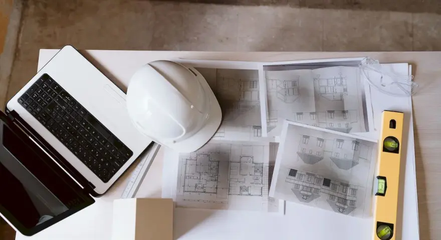 Laptop, kask, poziomica i plany architektoniczne – kompletny zestaw do pracy nad projektem technicznym.