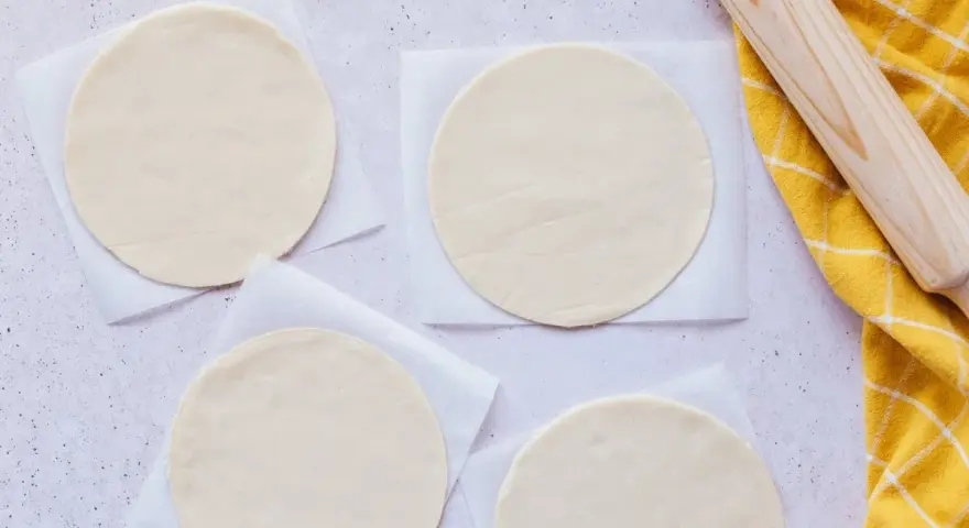 Cuatro discos de masa empanada listos para rellenar, junto a un rodillo y un paño amarillo.