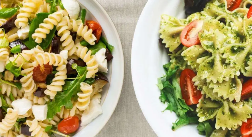 Due ciotole di pasta fredda gourmet: una con fusilli, melanzane, pomodorini e mozzarella; l'altra con farfalle al pesto e pomodorini.