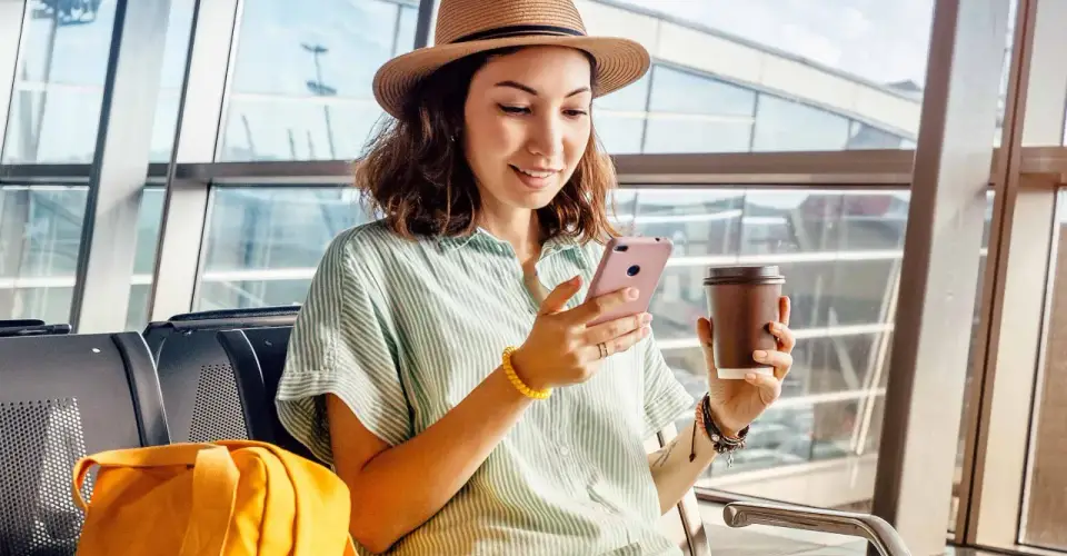 Kobieta w kapeluszu siedzi w poczekalni lotniskowej, trzymając telefon i kawę.