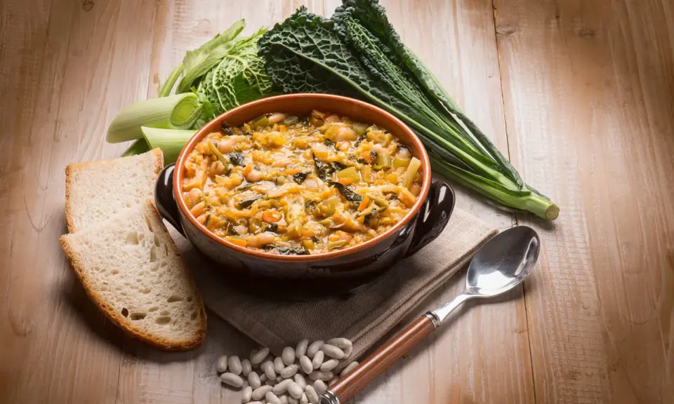 Piatto tipico toscano con pane raffermo: ribollita fumante in ciotola, contornata da fagioli, cavolo nero e porri.