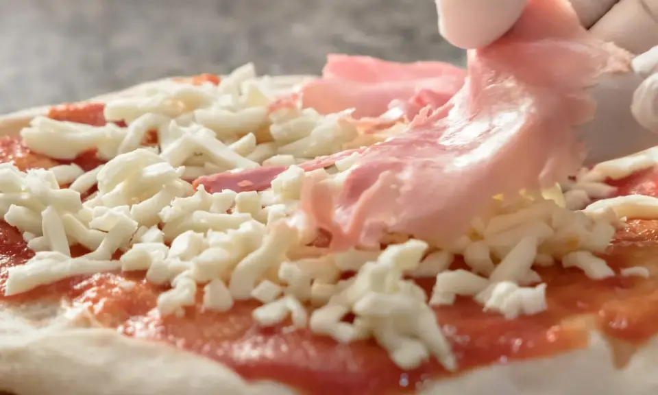 Rękawiczki zakładają cienkie plastry szynki parmeńskiej na pizzę z serem i sosem pomidorowym.