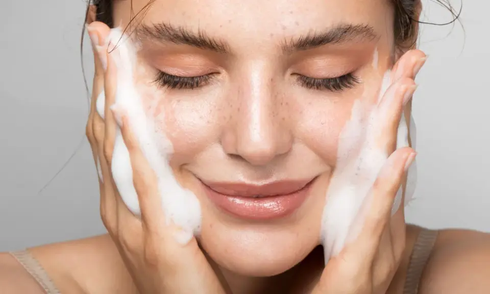 Donna con gli occhi chiusi mentre si lava il viso con un detergente schiumoso. Un gesto delicato per capire come si usa il detergente viso.