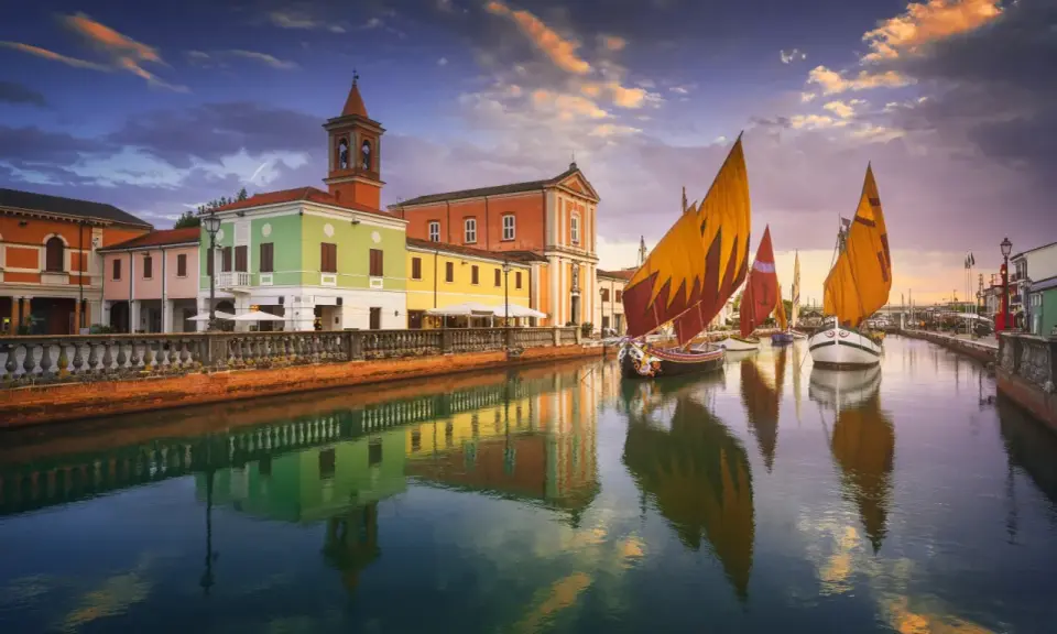 Navi a vela colorate navigano sul canale di Cesenatico al tramonto, con edifici vivaci e riflessi sull'acqua. Meglio Cervia o Cesenatico? Cesenatico offre questo spettacolo.