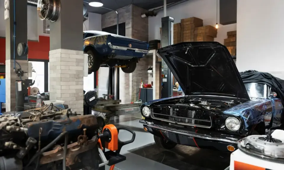 Un taller ordenado con un coche clásico negro con el capó abierto y otro azul en elevación. Motores y herramientas a la vista.