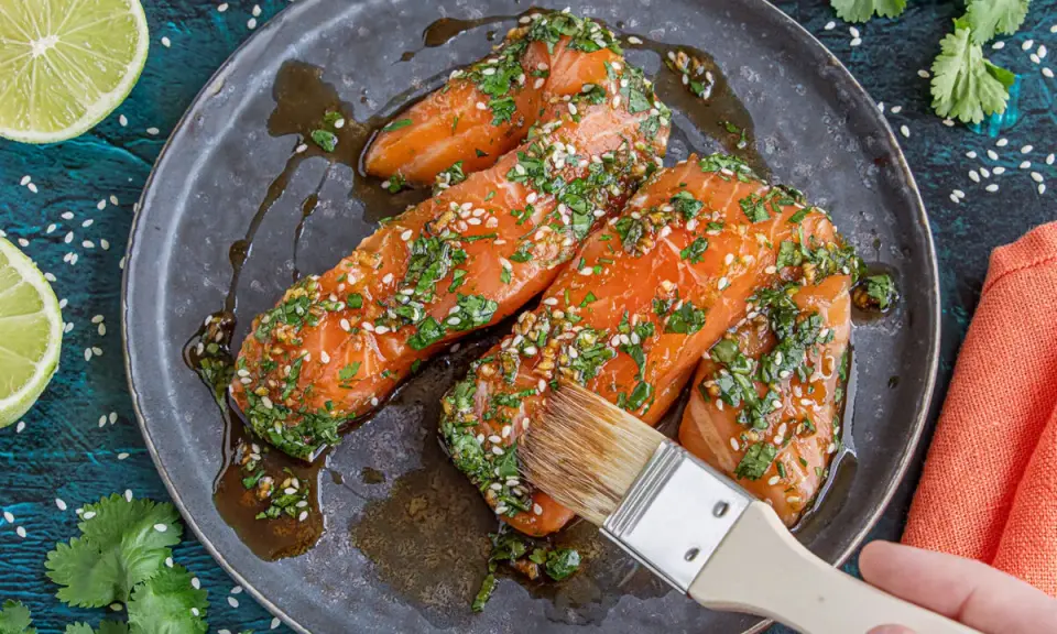 Vorbereitung für ein köstliches Gericht: Lachs marinieren mit Kräutern und Sesam. Eine Hand mit Pinsel verteilt die Marinade.