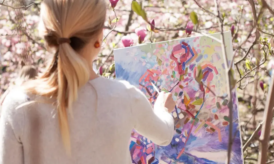En kvinna målar en abstrakt tavla utomhus bland blommande magnoliaträd. Hon använder olika målartekniker för att fånga vårens färger.