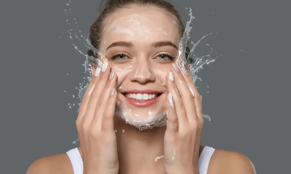 Mujer sonriente con espuma y agua en la cara, disfrutando de su rutina de doble limpieza.