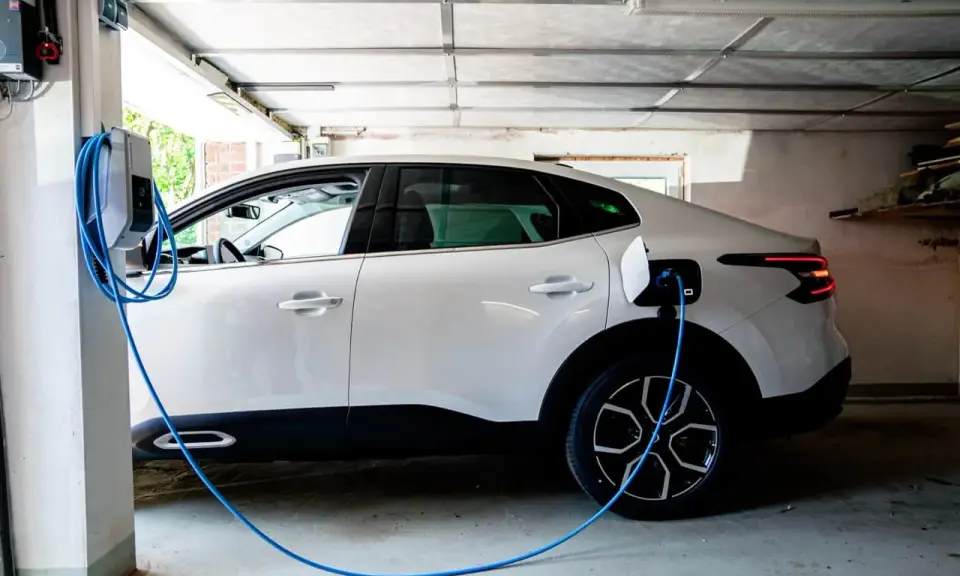 Vit elbil laddas i garage. En blå laddningskabel går från väggen till bilen. Tänk på brandrisk vid laddning av elbil i garage.