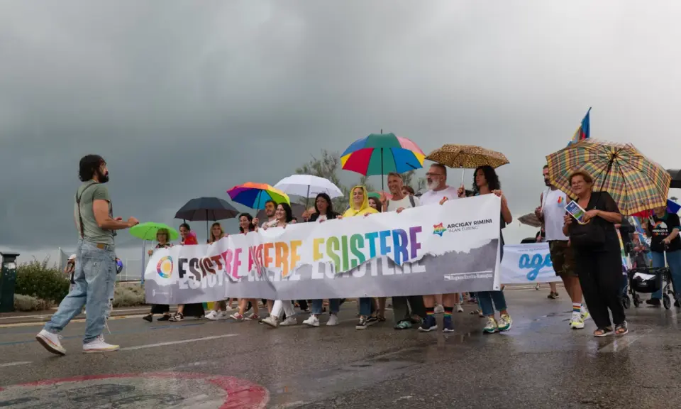 Persone con ombrelli colorati sfilano a Rimini Pride, tenendo uno striscione arcobaleno con scritto "Esistere Mere Esistere".