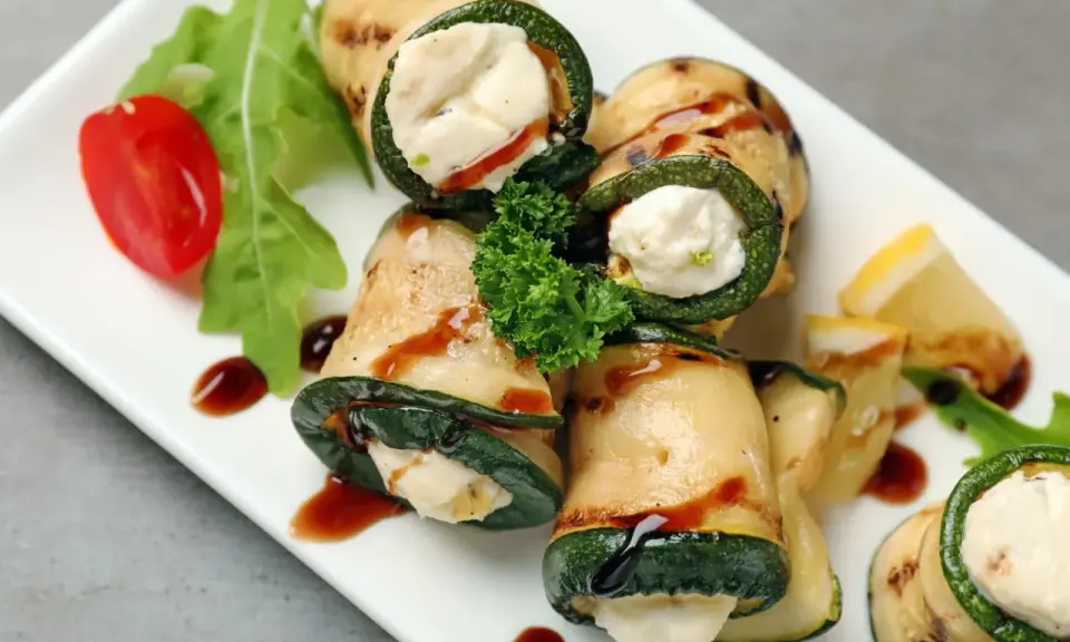 Deliziosi involtini di zucchine grigliate ripieni di formaggio cremoso, guarniti con pomodorini, rucola e salsa scura.