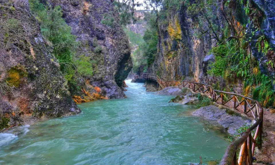 Un río turquesa serpentea entre cañones rocosos, con un sendero de madera que acompaña el recorrido. Ideales para rutas senderismo Jaén.