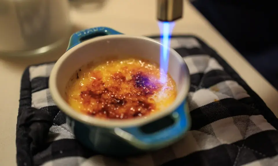 Flamme bleue caramélisant le sucre d'une crème brûlée, rappelant son origine.