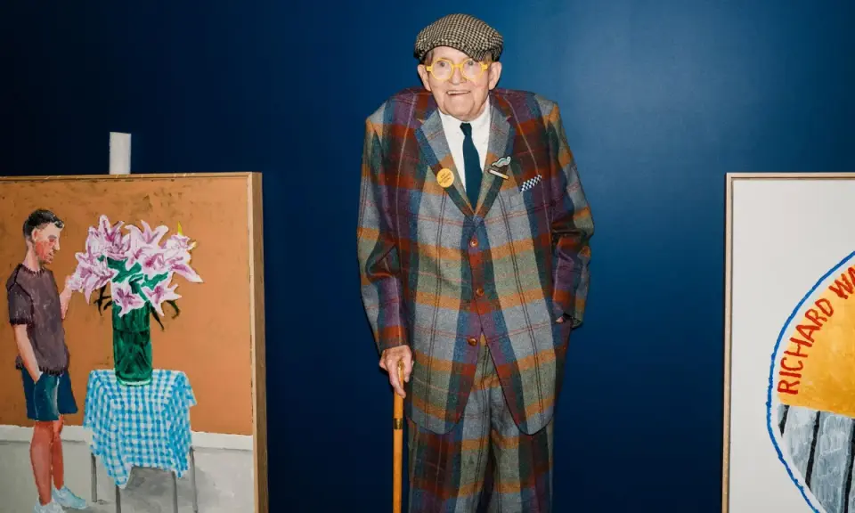 Ein Mann im karierten Anzug, inspiriert von David Hockney Pop Art, steht neben seinen lebendigen Gemälden.