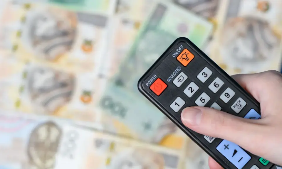 Dłoń trzyma pilota na tle polskich banknotów. Czy abonament RTV i zameldowanie mają związek z kosztami?