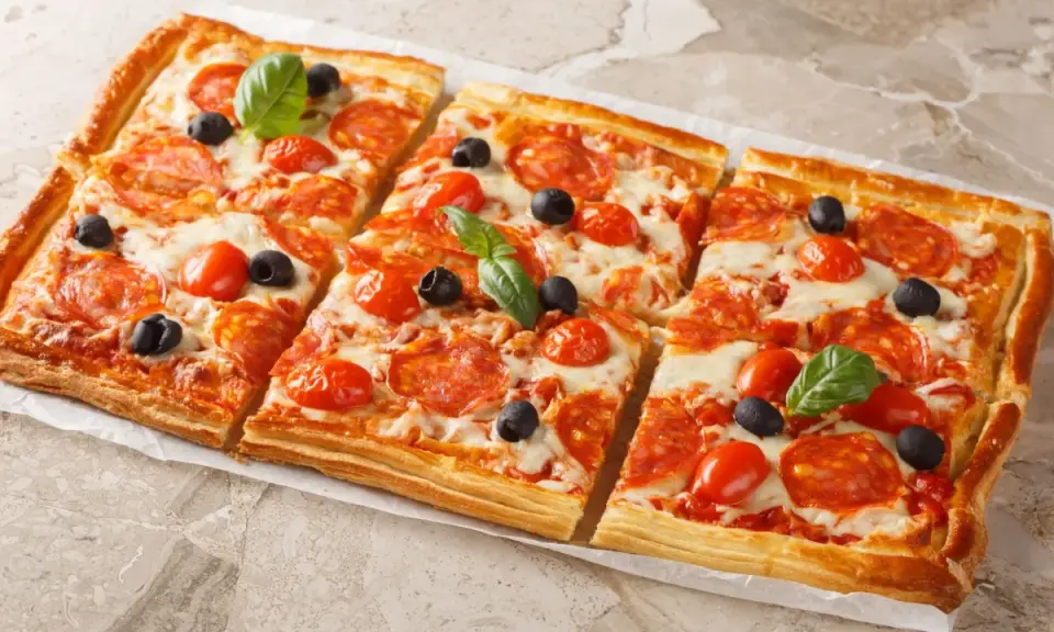 Chrupiąca pizza z ciasta francuskiego z salami, pomidorkami i oliwkami, udekorowana listkami bazylii.