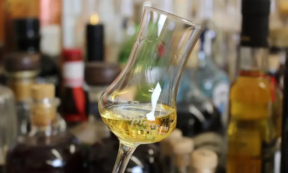 Ein Glas mit goldgelbem Wein steht vor einer Sammlung von Flaschen. Das Glas erinnert an ein Kirchenfenster, das Licht bricht.