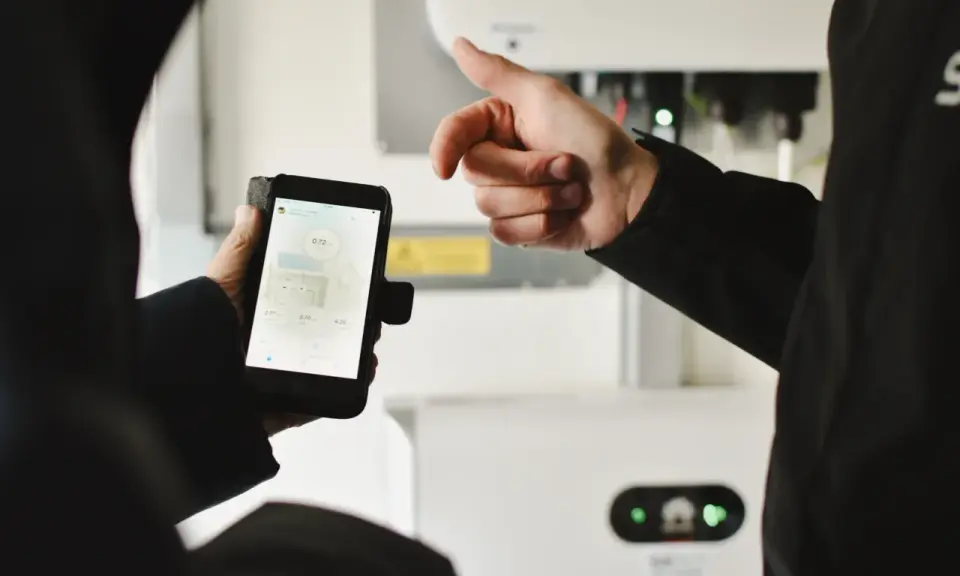 En person håller upp en mobiltelefon med sesol appen som visar solenergi-data. En annan person pekar mot en vit enhet i bakgrunden.