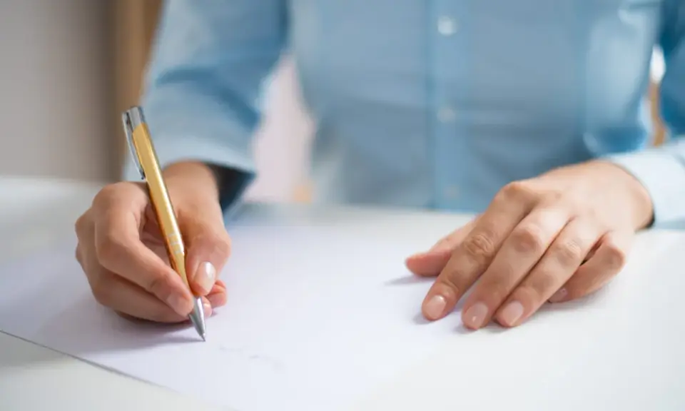 Dłoń pisze złotym długopisem na białej kartce, przygotowując się do egzaminu CAE practice tests.