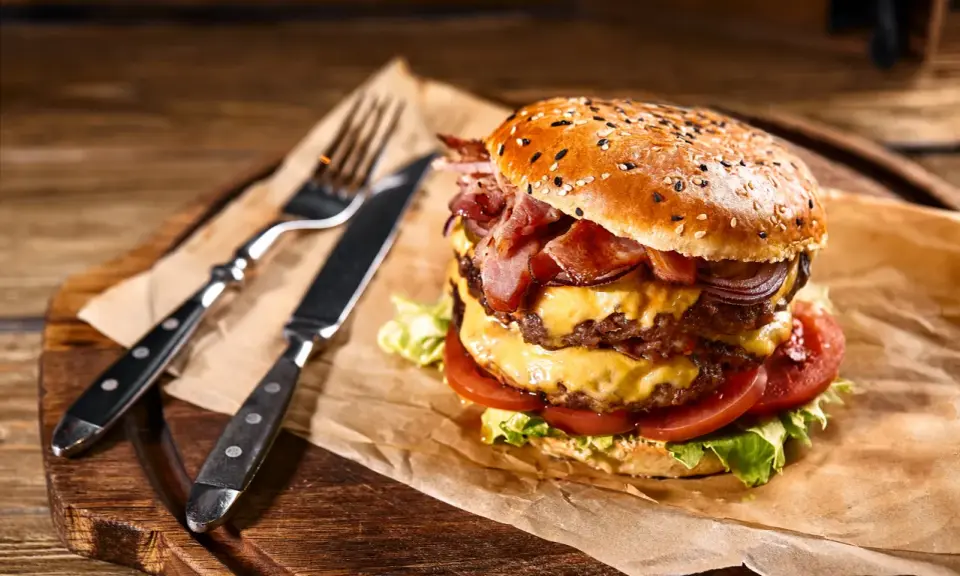 Soczysty burger z podwójną porcją mięsa, serem, bekonem i warzywami na desce z widelcem i nożem.