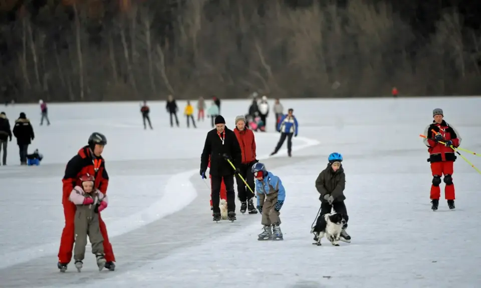 Människor åker norrviken skridskor på en isbelagd sjö. En pappa hjälper sitt barn, en hund dras på isen, och andra njuter av vintersporten.