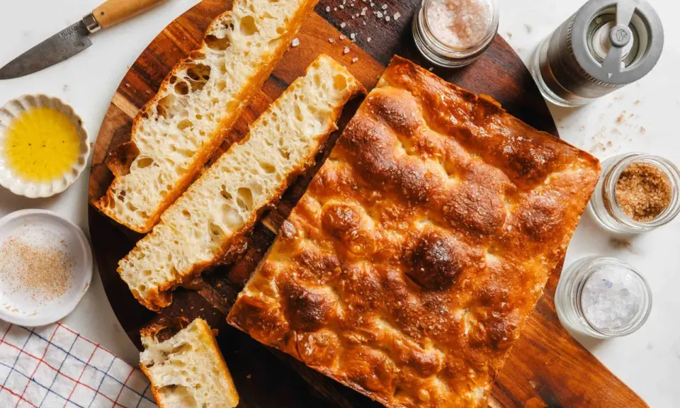 Goldbraune Focaccia mit grobem Salz, bereit zum Belegen. Daneben Olivenöl, Gewürze und ein Messer.