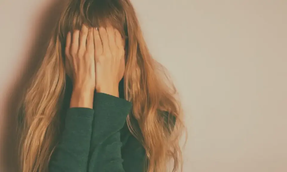 Kobieta z długimi blond włosami zakrywa twarz dłońmi na tle jasnej ściany.