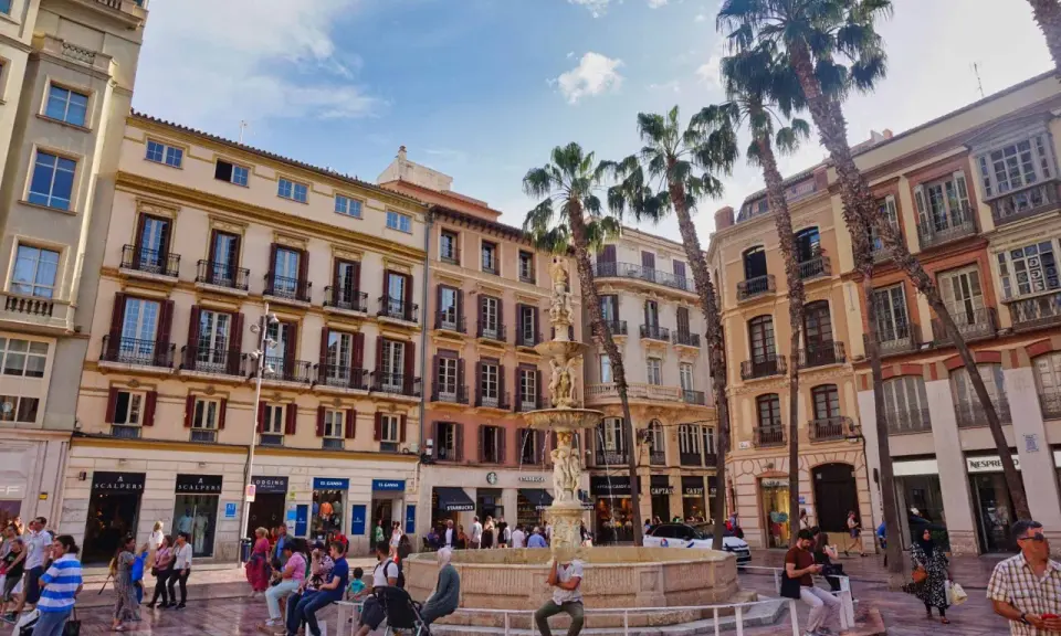 Sonniger Platz in der **Malaga Altstadt** mit einem Brunnen, Palmen und bunten Gebäuden. Menschen flanieren und genießen die Atmosphäre.