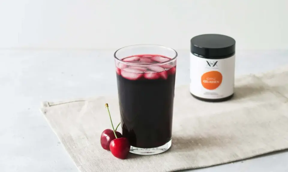 Ein Glas dunkler Sauerkirschsaft mit Eiswürfeln und zwei Kirschen daneben. Eine Dose mit der Aufschrift "Ganz Gelassen" steht im Hintergrund. Die positive sauerkirschsaft wirkung wird hier visuell dargestellt.