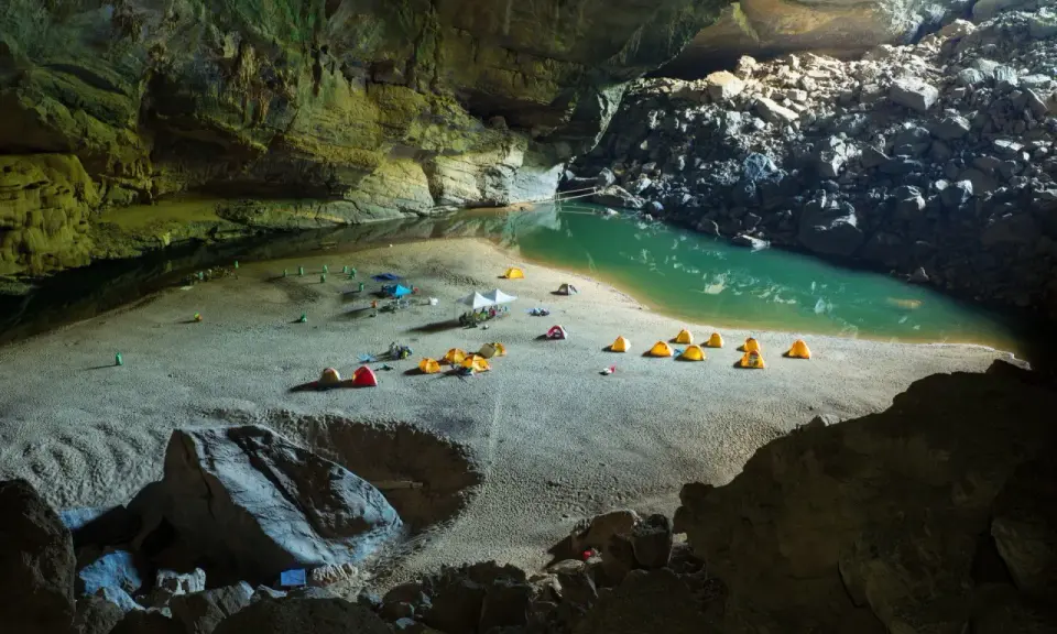 Obóz w jaskini Son Doong: namioty rozbite na piaszczystej plaży nad turkusową wodą, otoczone skalnymi ścianami.