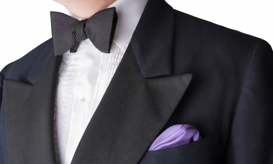 A man wears a black tuxedo with a pleated white shirt and a black bow tie. A purple pocket square adds a pop of color. This outfit answers the question: can you wear a tie with a tux? (No, a bow tie is traditional.)