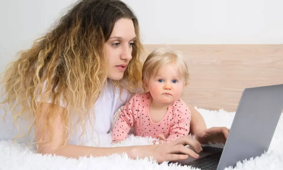 Kobieta z dzieckiem na rękach pracuje na laptopie, zastanawiając się, czy na urlopie wychowawczym można pracować.