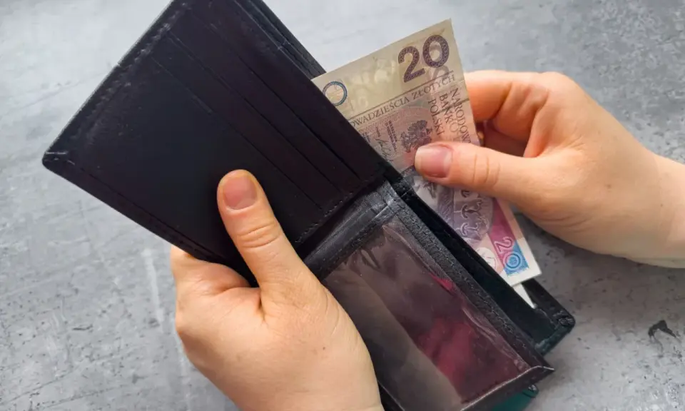 Ręce wkładają banknot 20 zł do portfela. Dowiedz się, komu przysługuje zasiłek dla bezrobotnych.