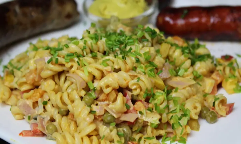 Ein cremiger nudelsalat mit mayo, garniert mit Schnittlauch. Daneben liegen zwei gebratene Würstchen und ein Schälchen Senf.