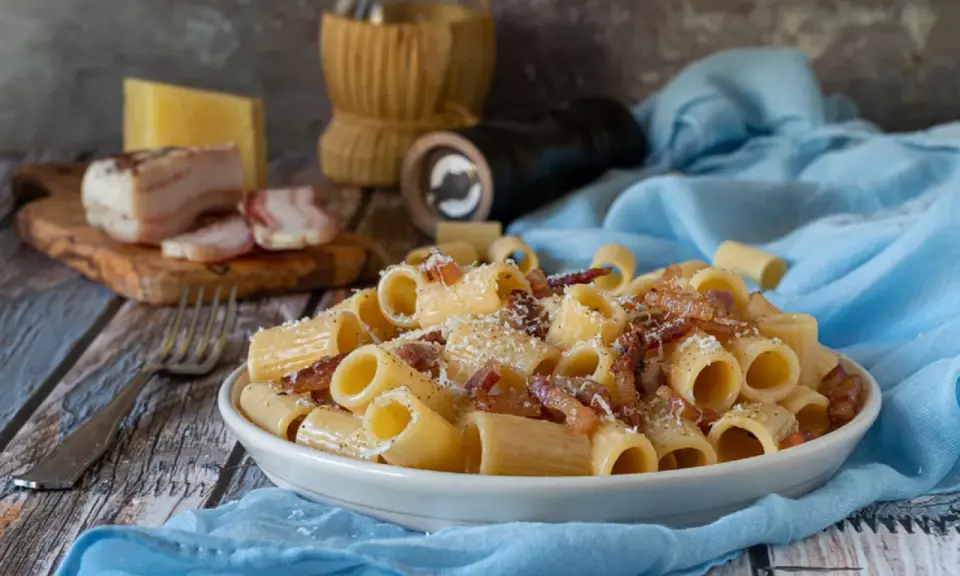Piatto di pasta alla gricia fumante, guanciale croccante e pecorino romano grattugiato. Ingredienti freschi su un tavolo rustico.