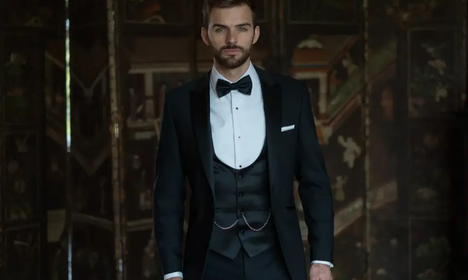 A dapper man in a black suit wedding guest attire, complete with a bow tie and waistcoat, stands confidently.