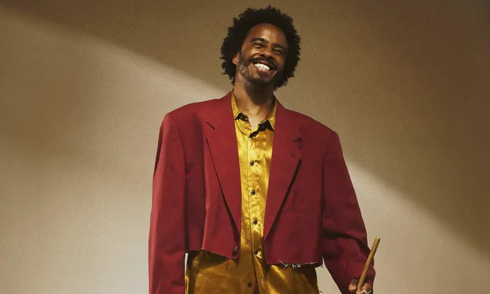 Un percussionniste célèbre, souriant, porte une veste courte bordeaux et une chemise dorée, tenant une baguette.
