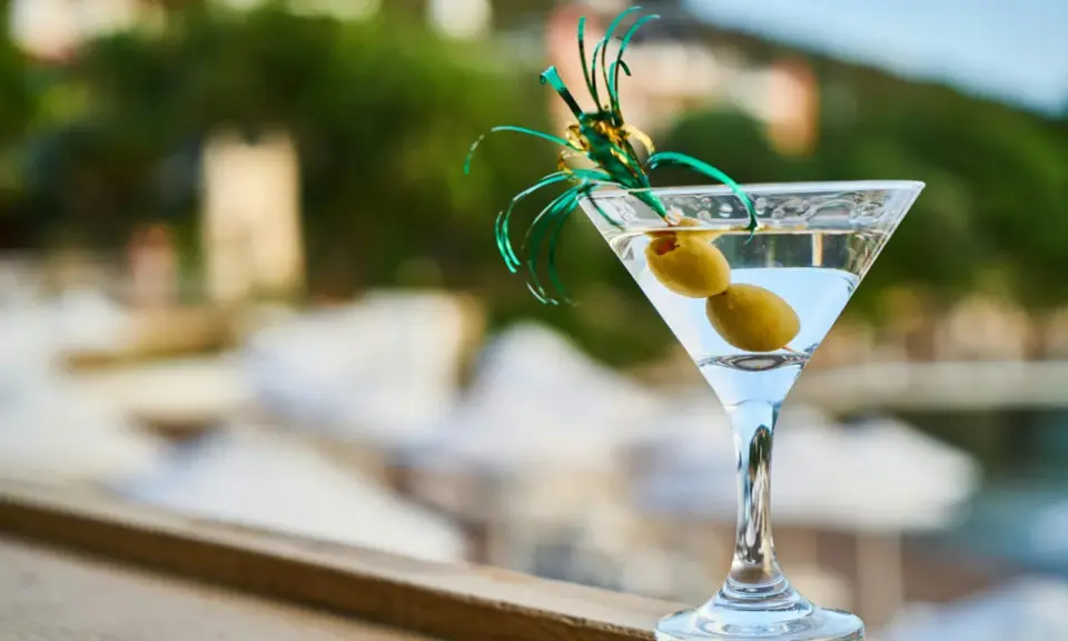 Idealny martini aperitivo z dwiema oliwkami, ozdobiony zielonymi wstążkami, na tle letniego krajobrazu.