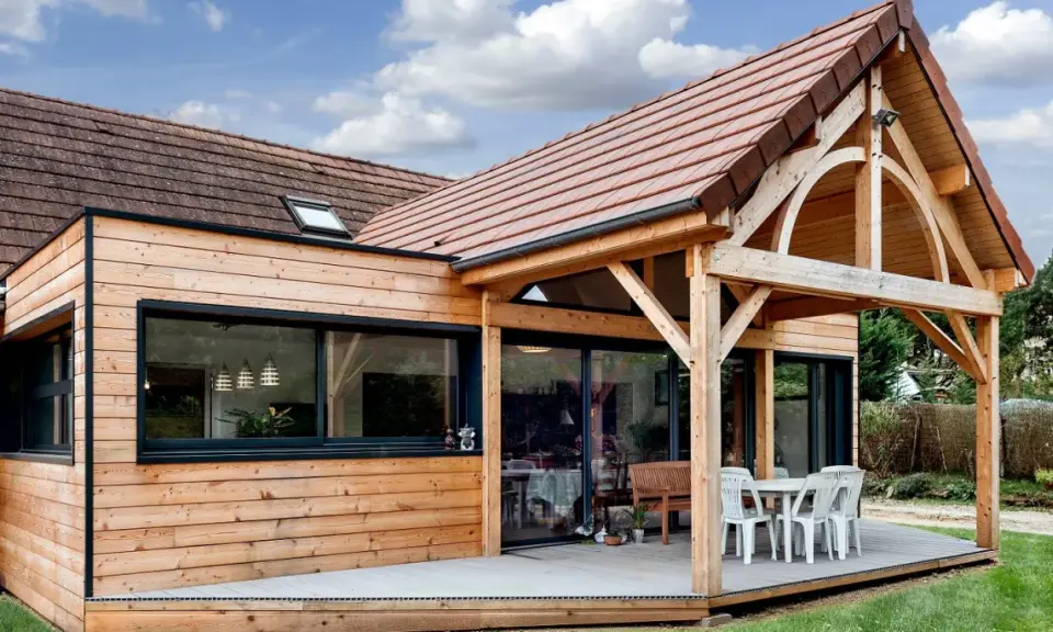 Extension de maison en bois avec terrasse sur dalle béton. Une table et des chaises sont installées sous un auvent.