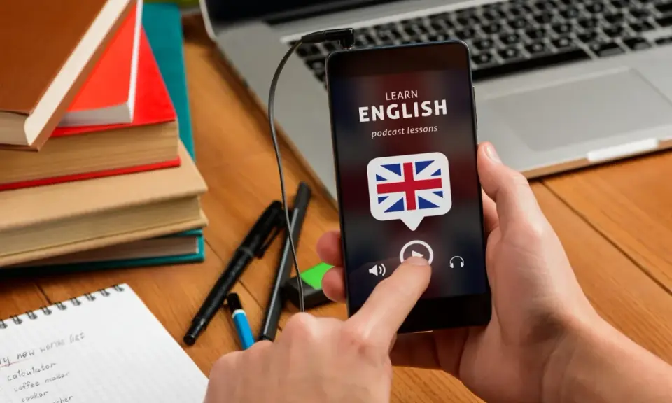 Ręka trzyma smartfon z aplikacją do nauki angielskiego. Na biurku leżą książki i notatnik z nowymi słówkami.