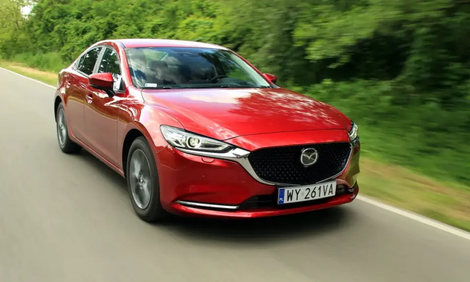 Czerwona Mazda 6 2.0 benzyna pędzi po wiejskiej drodze, otoczona zielenią drzew.