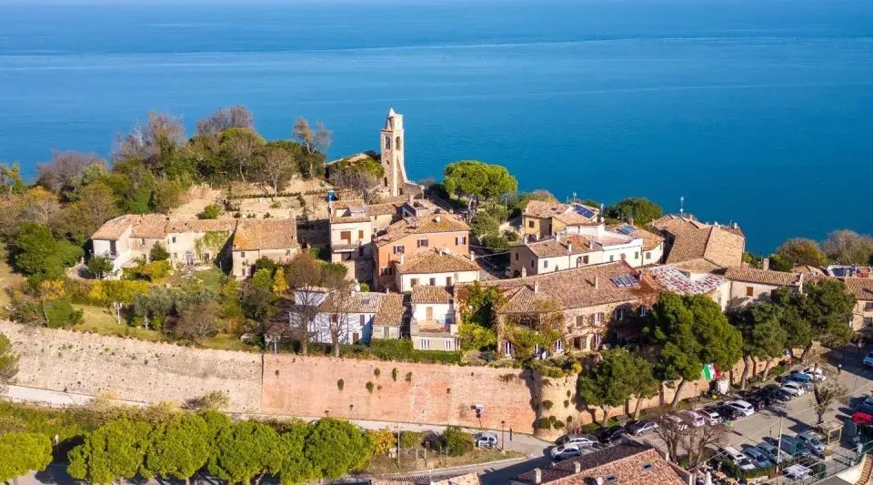 Borgo antico con campanile affacciato sul mare Adriatico. Il centro storico del lungomare Gabicce Mare, con tetti rossi e vegetazione rigogliosa, si estende verso l'azzurro intenso dell'acqua.