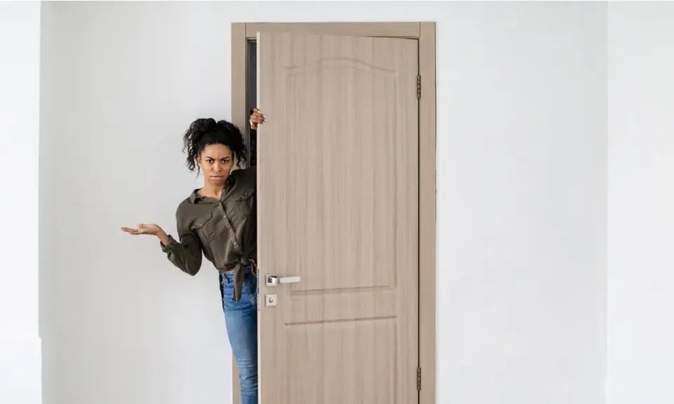 Mujer asomándose por una puerta de madera clara, con expresión de duda. Los colores de puertas como esta son muy versátiles.