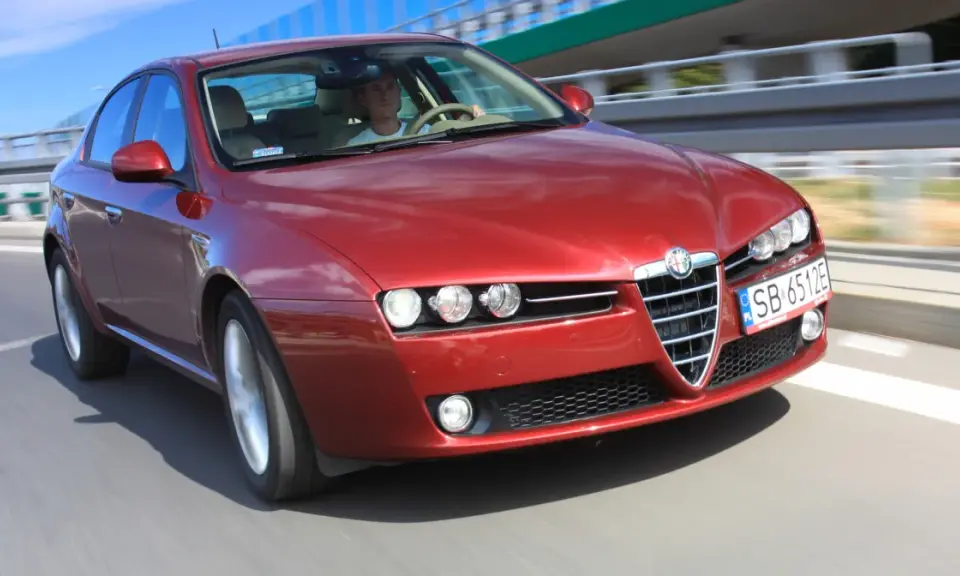 Czerwone Alfa Romeo 159 1.9 JTD pędzi po drodze, kierowca skupiony na trasie.