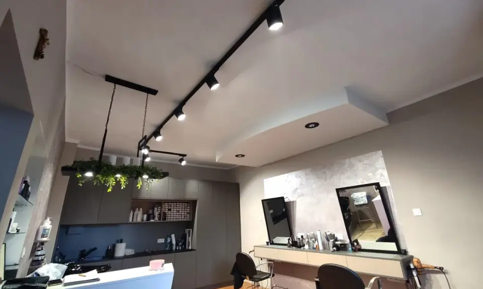 Nowoczesne oświetlenie w salonie fryzjerskim tworzy przytulną atmosferę. Reflektory na szynach i wisząca lampa z zielenią podkreślają styl wnętrza.