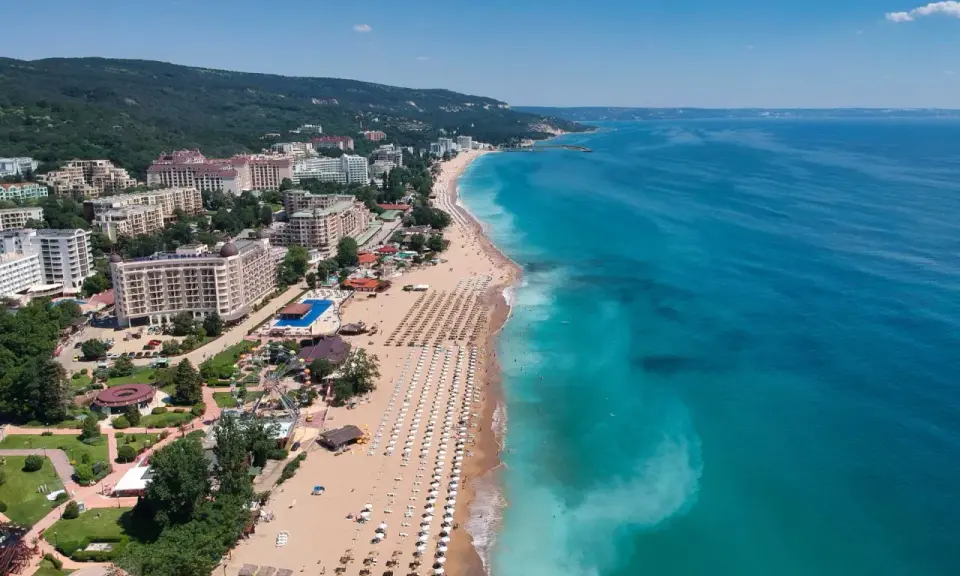 Słoneczny dzień nad morzem, gdzie złote piaski plaży wylegują się w słońcu, a turkusowa woda zaprasza do kąpieli. W tle hotelowe budynki i zielone wzgórza.